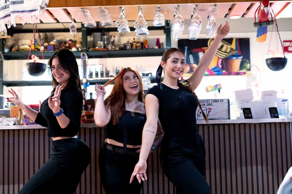 Tres meseras de Las Palmas sonriendo en la barra, bajo las copas, junto al letrero que dice que los clientes valen oro.